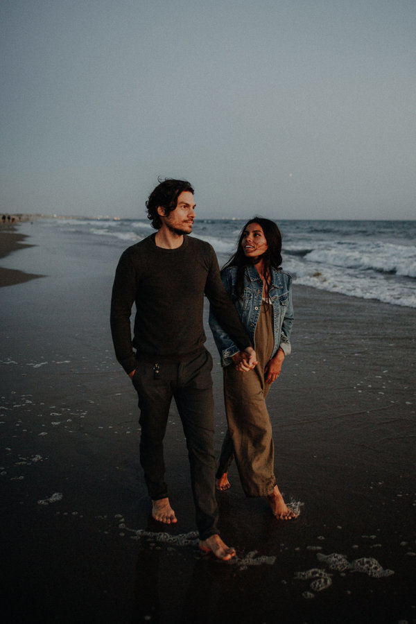 Auf dem Bild siehst du ein natürliches Foto von einem Paar, dass am Strand entlang läuft. Der Mann schaut auf das Meer und seine Freundin sieht ihn liebevoll an. Alles wirkt sehr natürlich.